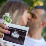 Expecting couple holding ultrasound image, celebrating pregnancy outdoors with a kiss.