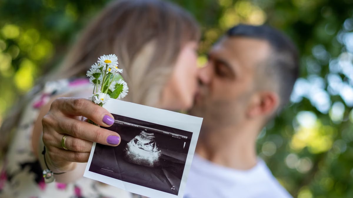 Expecting couple holding ultrasound image, celebrating pregnancy outdoors with a kiss.