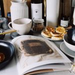 One portion of homemade cheese pancakes in frying pan and another on plate locating near opened stylish magazine among kitchen utensils and appliances in bright kitchen at home