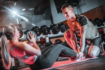A personal trainer assists a woman doing sit-ups with a medicine ball in a gym setting.